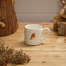 A white mug with a robin design sits on a wooden table. It's surrounded by rustic wood and pinkish dried flowers, creating a cosy, rustic vibe.