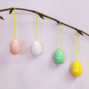 Four pastel egg hanging decorations with white polka dots – pink, white, green and yellow – suspended on yellow string from a bare branch against a pale pink background.