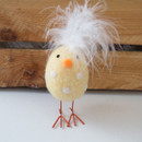 Yellow woollen chick decoration with white polka dots, fluffy feather tuft, orange beak and wire legs, standing in front of a wooden background.