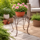Terracotta plant pot with pink flowers displayed on a brown metal plant stand on a patio, with outdoor seating and greenery in the background.