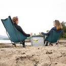 Two people sit on a sandy beach in teal chairs, facing each other. A cooler is between them. The scene is peaceful, with a distant shoreline and cloudy sky.