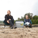 Two women sit on foldable chairs on a sandy beach, smiling and relaxed. A cooler is between them, with trees and a castle ruin in the background.
