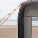 Close-up of an inflatable tent corner with taut orange ropes against a sandy beach and blurred ocean horizon, conveying a sense of outdoor adventure.