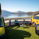 Three people sit on camping chairs by a lake, surrounded by vans and a fabric windbreak. They're enjoying a sunny day under clear blue skies.