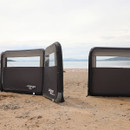 Inflatable windbreaks set up on a sandy beach, with the calm sea and distant mountains in the background. The scene is peaceful and serene.