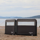Two black Vango Airbeam inflatable windbreak panels on a beach, with mountains and a calm sea in the background. The scene is peaceful and serene.