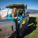 A woman in a turquoise jacket assembles a windbreak attached to a yellow camper van by a lake. The scene is sunny, with a clear blue sky.