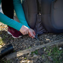 Person in a teal long-sleeve shirt secures a grey inflatable with a tent stake on rocky ground. They wear maroon sneakers, suggesting an outdoor setting.