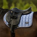 A black saddle rests on a blue, quilted LeMieux saddle pad on a brown horse. The background is a blurred mix of green hues, evoking calmness.