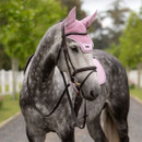 A dappled grey horse stands on a path wearing pink ear covers and saddle pad. The setting is a tree-lined avenue with a calm, serene atmosphere.