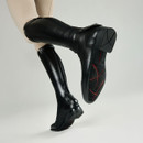 Close-up of a person wearing black equestrian boots with beige riding pants. The boots have textured soles with red accents, suggesting durability.