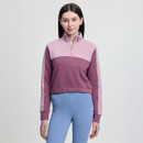 A woman wearing a mauve and pink quarter-zip pullover and blue leggings stands against a plain background. She has long dark hair and a subtle smile.