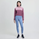 A woman stands smiling in a studio, wearing a purple and pink zip-up sweatshirt, light blue leggings, and navy sneakers. The background is plain white.