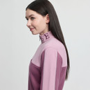 Young woman with long dark hair in profile, wearing a two-tone pink top with a high collar featuring embroidered letters. She has a slight smile.