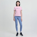 A woman stands against a plain background wearing a pink T-shirt and blue leggings. She smiles slightly, conveying a relaxed mood. Navy sneakers and white socks with navy stripes complete her outfit.