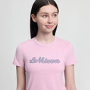 Young woman with straight brown hair, wearing a light pink T-shirt with "LeMieux" text. She gazes to the side with a soft smile, against a plain background.