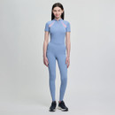 A woman stands against a plain light gray background wearing a fitted light blue athletic jumpsuit with pink accents, paired with black sneakers, conveying a sporty and confident tone.