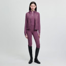 Woman in a matching mauve equestrian outfit, including a vest and long-sleeve top, stands confidently. She wears tall black boots, set against a light gray background.