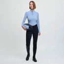 A smiling person holds a helmet, wearing a light blue zip-up top, black riding pants, and tall boots, set against a plain background. The mood is confident.