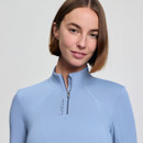 A smiling person holds a helmet, wearing a light blue zip-up top, black riding pants, and tall boots, set against a plain background. The mood is confident.