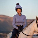 Smiling person wearing a light purple riding outfit and helmet sits on a white horse with braided mane against a soft, mountainous backdrop at sunset.