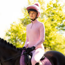 A person in a pink equestrian outfit and helmet rides a dark horse with a matching pink saddle pad. Sunlight filters through green trees, creating a serene atmosphere.