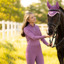 A woman in a purple outfit gently holds a black horse's bridle in a sunny field. The horse wears a matching purple veil, conveying harmony and calm.