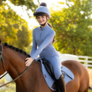 Young rider in blue equestrian attire smiles while seated on a brown horse with a matching saddle. Sunlit trees create a serene, cheerful backdrop.