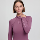 Woman with a bob haircut wearing a mauve LeMieux zip-up top, smiling softly. Her hand rests on her neck, against a neutral background.