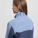 A woman with long hair, viewed from behind, wears a blue colour-block jacket with "MI" embroidered on the collar. The setting is minimal and neutral.