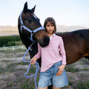 A woman in a pink shirt and denim shorts stands confidently beside a brown horse, holding a blue lead rope. They are outdoors, with mountains in the background.