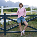 A woman stands against a dark wooden fence in a scenic field with mountains in the background. She wears a pink long-sleeve shirt and denim shorts, looking relaxed.