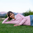 A woman in a pink sweater with "LEMIEUX" lies on grass, smiling warmly. Mountains and greenery form the calm backdrop, conveying a relaxed atmosphere.