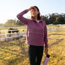 A woman wearing a mauve jacket smiles warmly while shielding her eyes from the sun. She stands near a paddock with horses and holds a water bottle.