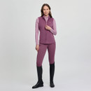 A woman stands confidently against a plain light grey background, wearing a mauve athletic vest and leggings with black riding boots. She exudes a calm, poised demeanour.
