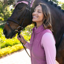 A woman in a pink vest smiles contentedly beside a brown horse with braided mane and pink ear net. The setting is a sunny, green garden path.