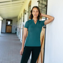 A woman in a teal athletic shirt stands in a stable corridor, resting her hand on her head. The setting is bright and airy, with wooden stable doors.