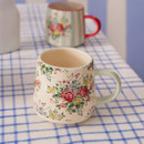 Cream mug with a sage green handle and vintage-style floral print, on a blue checked tablecloth with another mug in the background.
