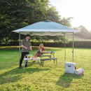 Couple enjoying a picnic under a Coleman pop-up gazebo canopy in a sunny garden, with folding table, benches and cool box on the grass.