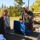 Two men at the back of a pickup truck opening a blue Coleman Snap ’N’ Go cooler outdoors.