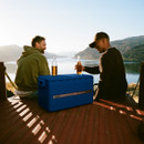 Two men sitting on a wooden platform overlooking a lake, using a closed Coleman Snap ’N’ Go cooler as a table for bottled drinks.