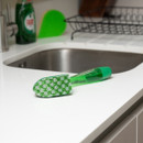 Green soap-dispensing dish brush with a patterned sponge head resting on a white kitchen countertop beside a sink, with washing-up liquid and a sponge blurred in the background.