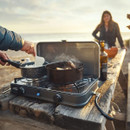 Portable two-burner gas camping stove set up on a wooden picnic table at the beach, with pots cooking on both burners and a person stirring food, while another person stands in the background near the shoreline at sunset.