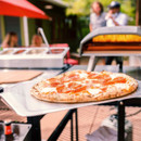 Pepperoni pizza on an Ooni pizza peel in front of a hot outdoor pizza oven during cooking.