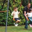 Child swinging on an orange seat while an adult pushes another child on the Hedstrom swing set.