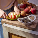 Person using a green apple corer and slicer to cut a red apple into even segments on a wooden worktop, with whole apples nearby.