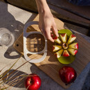 Hand placing neatly sliced apple segments on a green corer tray next to a white cutter tool on a wooden board, with whole apples around.