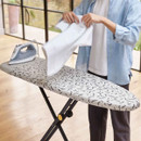Person folding a white garment on an ironing board with a patterned cover, with a steam iron resting to one side.