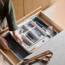 Person fitting an expandable cutlery organiser into a kitchen drawer alongside utensils and tools.