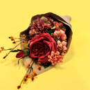 A bouquet of deep red and warm peach flowers lying on its side on a bright yellow background, partially wrapped in brown paper.